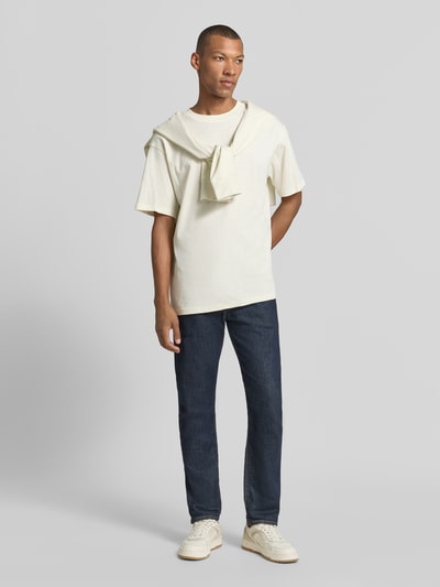 Männermodel trägt cremefarbenes Relaxed Fit T-Shirt aus reiner Baumwolle Modell 'MAARKOS' von ARMEDANGELS, kombiniert mit dunkler Jeans, Ganzkörperansicht von vorne.