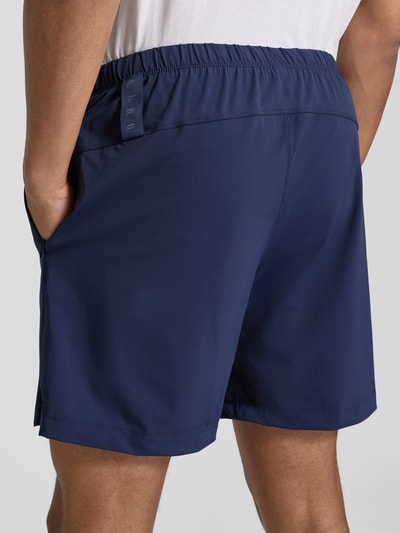 Mężczyznamodel nosi Regular Fit Shorts mit Logo-Print od Fabletics w granatowym kolorze, elastyczny materiał, szerokość w rozmiarze M: 39 cm, krój regular fit, elastyczny pas z tunelem, widok całej sylwetki z tyłu.