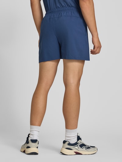 Männermodel trägt Shorts mit elastischem Bund und Logo von New Balance in Blau, Regular Fit, glatte Textur, Ganzkörperansicht Rückansicht.
