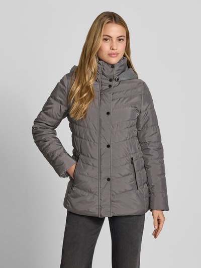 Modelka nosi 'Jacke in Stepp-Optik mit Kapuze' od FUCHS SCHMITT w kolorze szarym, z efektem pikowania, taliowany krój, kaptur, ocieplana stójka, wpuszczane kieszenie na zamek błyskawiczny, długość tyłu w rozmiarze 36: 74 cm, szerokość ramion w rozmiarze 36: 40 cm, szerokość klatki piersiowej w rozmiarze 36: 48 cm, długość rękawów w rozmiarze 36: 64 cm, ujęcie całej sylwetki od przodu.