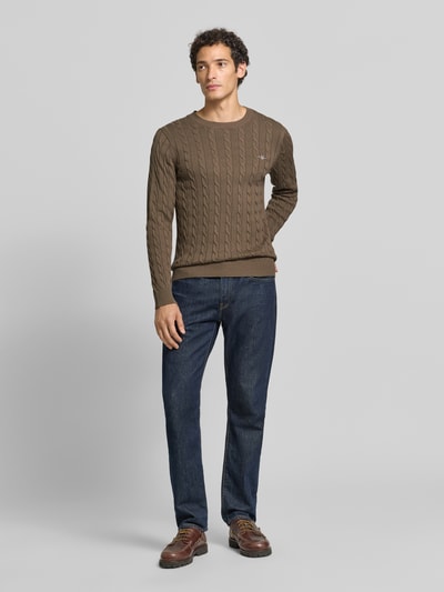 Männermodel trägt Regular Fit Pullover aus reiner Baumwolle von GANT in Braun mit Zopfmuster, kombiniert mit Jeans, Ganzkörperansicht von vorne.