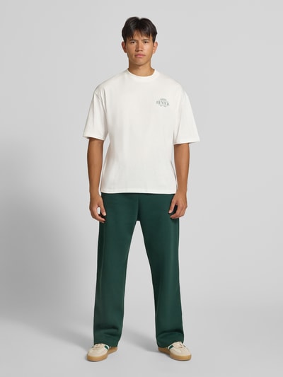 Männermodel trägt Regular Fit T-Shirt aus reiner Baumwolle mit rückseitigem Print von REVIEW, helles Shirt, grüne Hose, Ganzkörperansicht von vorne. Rückenlänge bei Größe M: 67 cm, Logo-Detail sichtbar.
