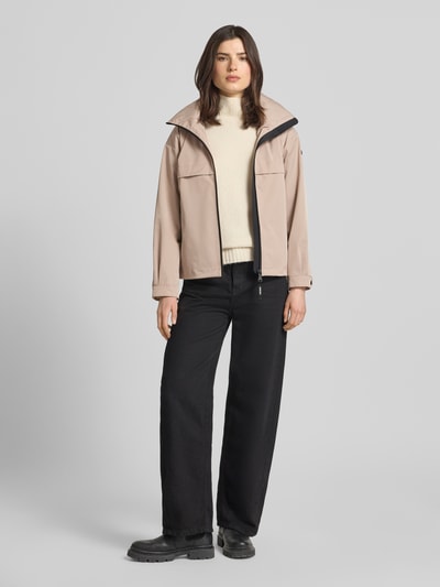 Kobiece modelka nosi kurtka z stójką Jacke mit Stehkragen od khujo, krój regular fit, zamek błyskawiczny, kieszenie zapinane na zamek błyskawiczny, długość tyłu w rozmiarze S: 60 cm, szerokość ramion w rozmiarze S: 51 cm, szerokość klatki piersiowej w rozmiarze S: 50 cm, długość rękawów w rozmiarze S: 54 cm, kolory beżowy i czarny, tekstura gładka, całkowity widok z przodu.