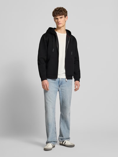 Männermodel trägt schwarze Sweatjacke mit Reißverschluss von Guess, katoenmix, los model, capuchon, tunnelkoord, steekzakken, ribboorden, ruglengte maat M 66 cm, mouwlengte maat M 66 cm, ganzkörperansicht von vorne