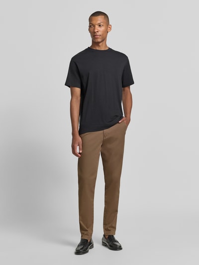 Männermodel trägt braune Slim Fit Chino aus Baumwoll-Mix Modell 'GABARDINE' von Selected, kombiniert mit schwarzem T-Shirt, Ganzkörperansicht von vorne. Herren Chino von Selected.