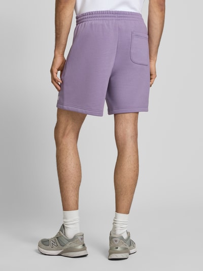 Männermodel trägt Relaxed Fit Shorts mit Logo-Stitching von New Balance in Flieder, Baumwoll-Mix, elastischer Bund, Ganzkörperansicht Rückansicht.
