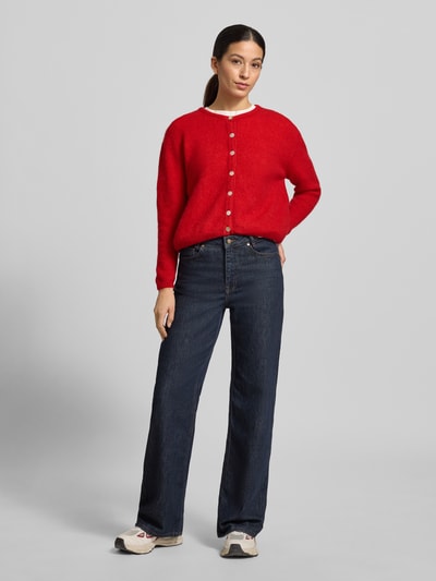 Frauenmodel trägt Strickjacke mit Knopfleiste Modell 'VITOW' von American Vintage, leuchtend rot, lockerer Schnitt, Jeans, Ganzkörperansicht von vorne.