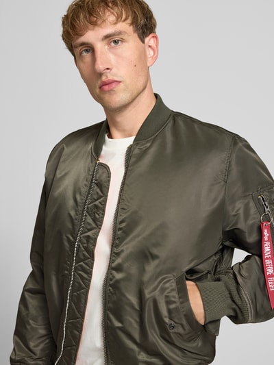 Männermodel trägt Bomberjacke mit Pattentaschen und Label-Applikation von Alpha Industries in Olivgrün, glatte Textur, Nahaufnahme Oberkörper. Regular Fit, Stehkragen, Reißverschluss, Maße: Rückenlänge 69 cm, Schulterbreite 44 cm, Brustweite 54 cm, Ärmellänge 64 cm.