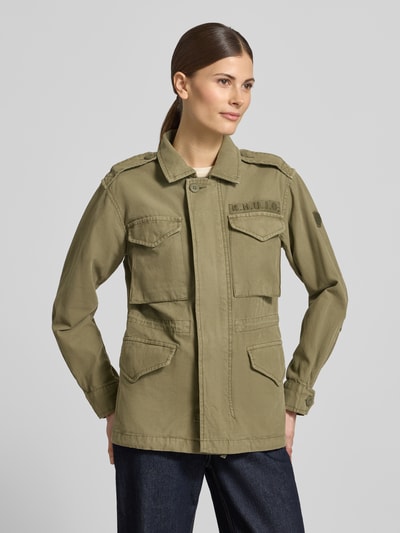 Modelka nosi kurtkę z bawełny o kroju regular fit, model 'Buni' od khujo, zielony kolor, nakładane kieszenie, wyhaftowane logo, wykładany kołnierz, długość tyłu w rozmiarze S: 60 cm, szerokość ramion w rozmiarze S: 40 cm, szerokość klatki piersiowej w rozmiarze S: 50 cm, długość rękawów w rozmiarze S: 56 cm. Ganzkörperansicht von vorne.