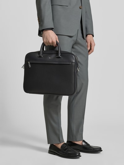 Männermodel trägt Laptoptasche in Lederoptik Modell 'RAY_MP_S DOC CASE' von BOSS in Schwarz mit Logo-Muster, Ganzkörperansicht von vorne.