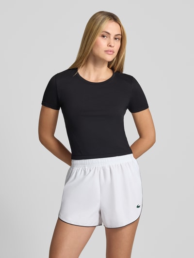 Kobieta modelka nosi damski t-shirt Cropped T-Shirt mit Rundhalsausschnitt Model 'Feather' od Fabletics, elastyczna mieszanka materiałów, krój krótki, okrągły dekolt, długość tyłu w rozmiarze S 45 cm, widok całej sylwetki z przodu.