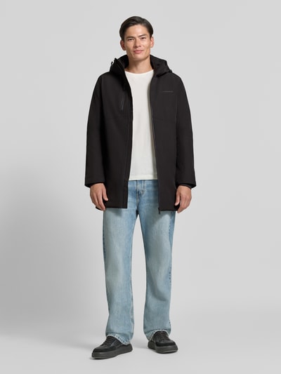 Męski model nosi Regular Fit Parka mit Logo-Print od Lindbergh, elastyczna mieszanka materiałów, czarna, kaptur z tunelem, napoleońska kieszeń, zamek błyskawiczny, Szerokość ramion w rozmiarze M 50 cm, Szerokość klatki piersiowej w rozmiarze M 58 cm, Długość tyłu w rozmiarze M 90 cm, Długość rękawów 65 cm, widok całej sylwetki od przodu.