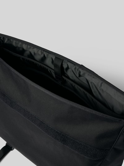 Produktfoto, Messenger Bag mit Label-Patch Modell 'Jake' von Carhartt Work In Progress, schwarzer Stoff, Innenansicht, Hauptfach sichtbar, Klettverschluss, glatte Textur, großes Laptopfach, minimalistisches Design