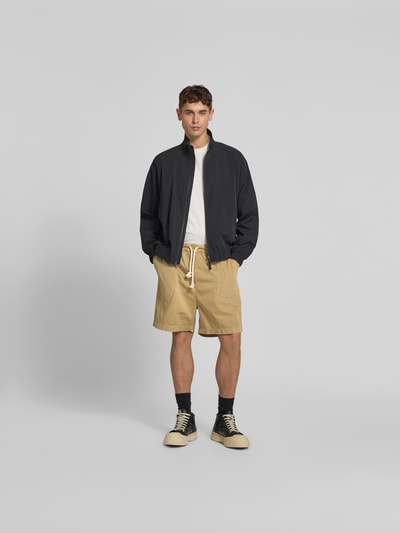 Männermodel trägt Bermudas aus reiner Baumwolle von Officine Générale, lockere Passform, elastischer Bund, Eingrifftaschen, Tunnelzug, Beige, Ganzkörperansicht von vorne.
