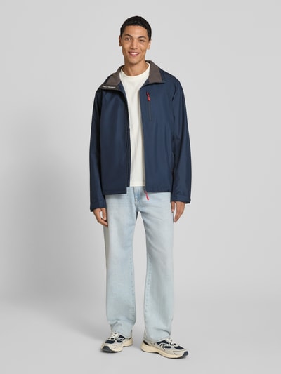 Männermodel trägt Regular Fit Jacke mit Stehkragen Modell 'Crew' von Helly Hansen, dunkelblau, Reißverschlusstaschen und Logo, Ganzkörperansicht von vorne.