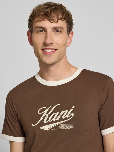 Męski model nosi T-Shirt mit Label-Print und Rundhalsausschnitt od Karl Kani, bawełna, krój regular fit, długość tyłu 68 cm, zbliżenie na górną część ciała.
