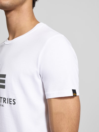 Męski model nosi białe T-Shirt mit Label-Print od Alpha Industries, czysta bawełna, prosty krój, okrągły dekolt, zbliżenie na górną część ciała.