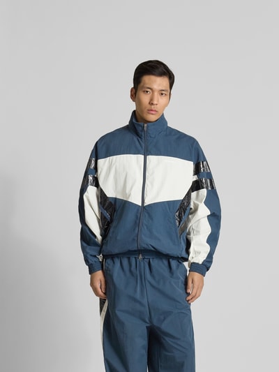 Männermodel trägt Trainingsjacke mit Stehkragen von VETEMENTS in Blau-Weiß mit schwarzen Details, elastische Bündchen, Relaxed Fit, Nahaufnahme Oberkörper.