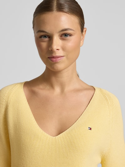 Kobiece modelka nosi Regular Fit Strickpullover aus reiner Baumwolle od Tommy Hilfiger w żółtym kolorze, V-dekolt, logo, prążkowany materiał, zbliżenie na górną część ciała.