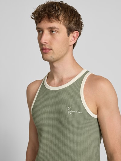 Męski model nosi Slim Fit Top mit Logo-Stitching od KARL KANI z czystej bawełny, krój slim fit, wzór strukturalny, okrągły dekolt, khaki z białymi lamówkami, długość tyłu w rozmiarze M: 68 cm, zbliżenie na górną część ciała.