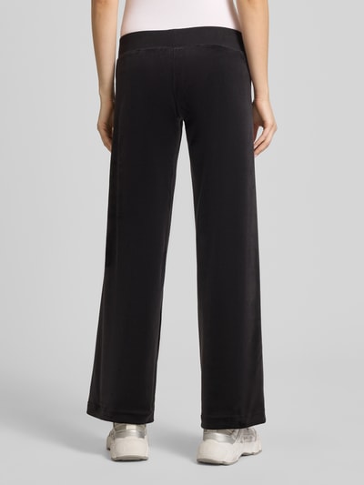 Frauenmodel trägt Sweatpants mit Ziersteinbesatz Modell 'GOTHIC CROWN' von Juicy Couture, czarny kolor, krój straight fit, materiał z dodatkiem stretchu, elastyczny pas ze sznurkiem, długość wewnętrzna nogawki w rozmiarze S 78 cm, widok całej sylwetki od tyłu.