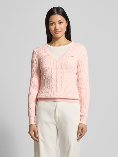 Modelka kobieca nosi różowy Strickpullover mit Zopfmuster und Label-Stitching od Gant, wzór warkoczowy, dekolt V, widok całej sylwetki z przodu.