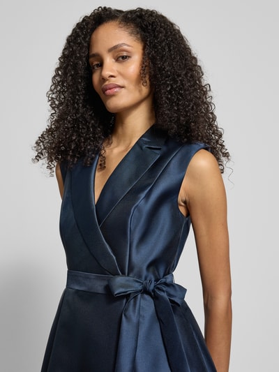 Frauenmodel trägt Cocktailkleid mit Reverskragen von Adrianna Papell, taillierter Schnitt, Taillengürtel, blickdichtes Unterkleid, Nahtreißverschluss, glänzender dunkelblauer Stoff, Ganzkörperansicht von vorne. Rückenlänge 38 cm, Brustweite 39 cm bei Größe 36.