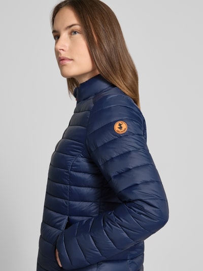 Frauenmodel trägt dunkelblaue Slim Fit Steppjacke mit gefüttertem Stehkragen Modell 'Carly' von SAVE THE DUCK, Nahaufnahme Oberkörper, Stepp-Optik, Zweiwegereißverschluss, Eingrifftaschen mit Reißverschluss, glatte Struktur.