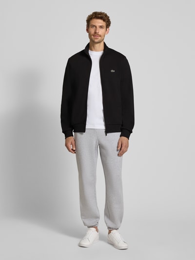 Männermodel trägt Regular Fit Sweatjacke aus Baumwoll-Mix von Lacoste in Schwarz, weiße Schuhe, graue Hose, Ganzkörperansicht von vorne. Herren Sweatjacke von Lacoste