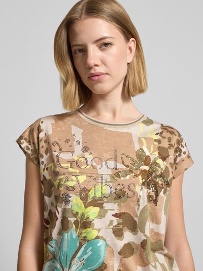 Frauenmodel trägt T-Shirt im Allover-Look mit Ziersteinbesatz von Betty Barclay aus Baumwoll-Modal-Mix, Regular-Fit, Rundhalsausschnitt, Blumenmotiv, Farben Beige, Grün und Türkis, Tunnelzug, Rückenlänge 57 cm bei Größe 36, Nahaufnahme Oberkörper.