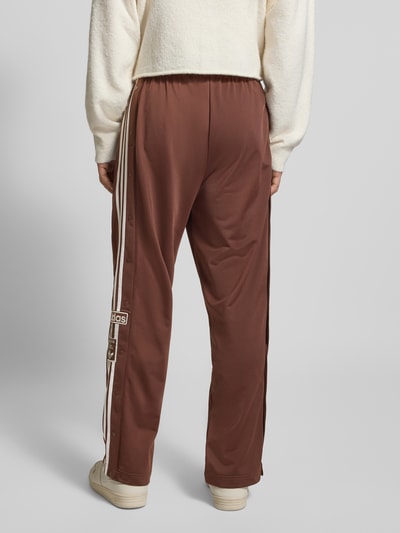 Männermodel trägt Wide Fit Jogginghose mit Logo-Stitching Modell 'Adibreak' von adidas Originals, braun, Knopfleisten, Logo-Patch, Ganzkörperansicht Rückansicht.