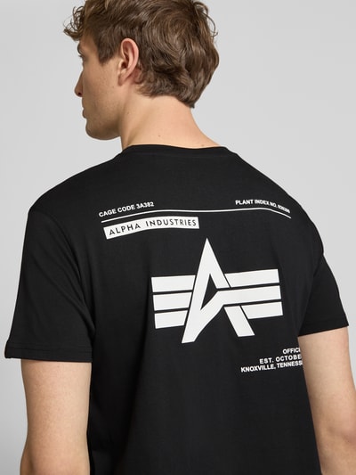 Männermodel trägt T-Shirt mit Label-Patch von Alpha Industries in Schwarz, große weiße Aufdrucke auf glatter Baumwolle, Rückenansicht, Reine Baumwolle, Gerader-Schnitt, Rundhalsausschnitt, Herren T-Shirt, Rückenlänge bei Größe M: 66 cm. Ganzkörperansicht Rückansicht.