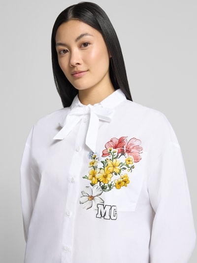 Frauenmodel trägt Relaxed Fit Hemdbluse aus reiner Baumwolle von Marc Cain in Weiß mit farbigem Blumenprint, Logo-Print und Stehkragen mit Tunnelzug, Nahaufnahme Oberkörper.