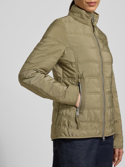 Modelka nosi kurtkę pikowaną z stójką i kieszeniami zapinanymi na zamek błyskawiczny, Steppjacke mit Stehkragen und Reißverschlusstaschen von Fuchs Schmitt, taliowany krój, detal z logo, zamek błyskawiczny dwustronny, długość tyłu w rozmiarze 36: 65 cm, szerokość ramion w rozmiarze 36: 45 cm, szerokość klatki piersiowej w rozmiarze 36: 42 cm, długość rękawów w rozmiarze 36: 65 cm, jednolity kolor oliwkowy, gładka faktura, Ganzkörperansicht z przodu.