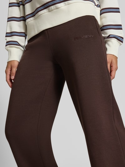 Frauenmodel trägt Sweathose mit geradem Bein von REVIEW in Braun, Logo-Print, Baumwoll-Mix, seitliche Taschen, Nahaufnahme Oberschenkel.