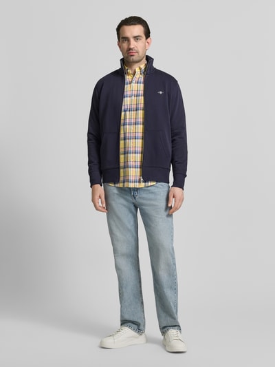 Męski model nosi Regular Fit Sweatjacke mit Stehkragen Modell 'Gield' od Gant, ciemnoniebieska bluza z bawełnianej mieszanki, logo, zamek, kieszenie, elastyczne brzegi, długość tyłu 69 cm, długość rękawa 64 cm, widok całej sylwetki z przodu.
