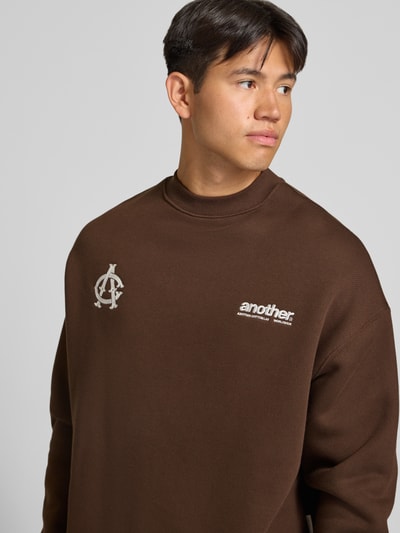 Męski model nosi bluza 'Sweatshirt mit Label-Stickerei' od ANOTHER COTTON LAB, mieszanka bawełny, luźny krój, okrągły dekolt, wyhaftowane logo, prążkowane wykończenia, kolor brązowy, tekstura gładka, zbliżenie na górną część ciała.