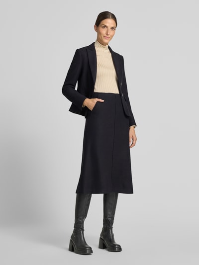 Frauenmodel trägt Slim Fit Kurzblazer aus reiner Schurwolle von Marc Cain in Schwarz mit Reverskragen, Knopfverschluss und Pattentaschen, kombiniert mit beigem Pullover und schwarzem Rock, glatte Stoffstruktur, Ganzkörperansicht von vorne. Rückenlänge 55 cm, Brustweite 33 cm, Ärmellänge 60 cm.