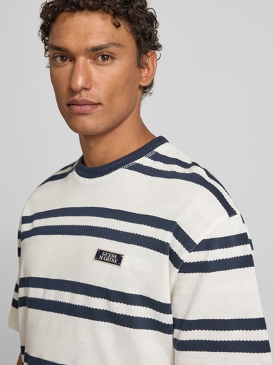 Männermodel trägt T-Shirt mit Label-Detail von Guess in Weiß und Marine mit Streifen, Reine Baumwolle, Lockerer Schnitt, Nahaufnahme Oberkörper.