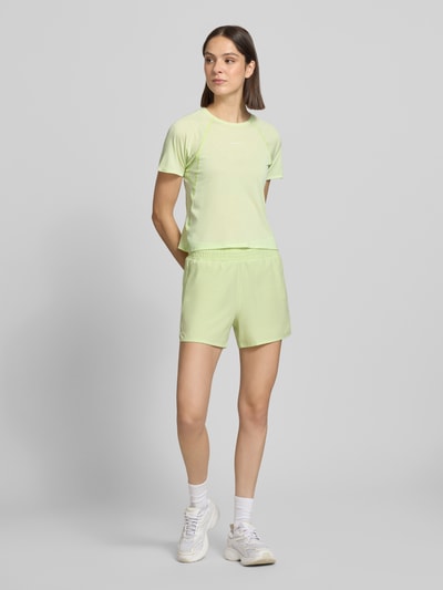 Frauenmodel trägt Salomon Damen T-Shirt Cropped T-Shirt mit Rundhalsausschnitt Modell 'SHKOUT' mit Raglanärmel und taillierter Passform in hellem Pastellgelb, dazu passende Shorts, glatte Textur, Rückenlänge bei Größe S: 47 cm, Ganzkörperansicht von vorne.