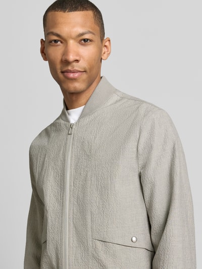 Männermodel trägt Bomberjacke mit Pattentaschen und Reißverschluss von PAUL SMITH, hellgrau, strukturierte Oberfläche, Nahaufnahme Oberkörper, Herren, Loose-Fit, Stehkragen, Reißverschluss, Druckknopfverschluss, gerippte Abschlüsse, Rückenlänge bei Größe M 65 cm, Schulterbreite 44 cm, Brustweite 53 cm, Ärmellänge 67 cm.