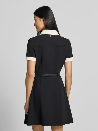 Frauenmodel trägt Minikleid mit Polokragen Modell 'Claudie' von GUESS, Schwarz mit weißen Details, strukturierter Stoff, Rückenansicht, Ganzkörperansicht Rückansicht. Taillierte Passform, kurzer Schnitt, elastischer Material-Mix, Brusttaschen, integrierter Gürtel, Knopfleiste, kurzärmelig.