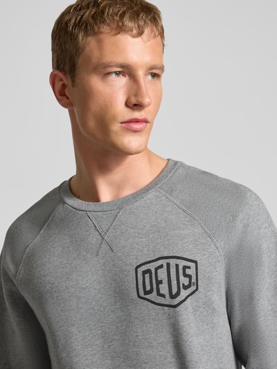 Männermodel trägt Sweatshirt mit Logo und Rundhalsausschnitt von DEUS EX MACHINA aus reiner Baumwolle in Grau, Relaxed-Fit, Nahaufnahme Oberkörper.