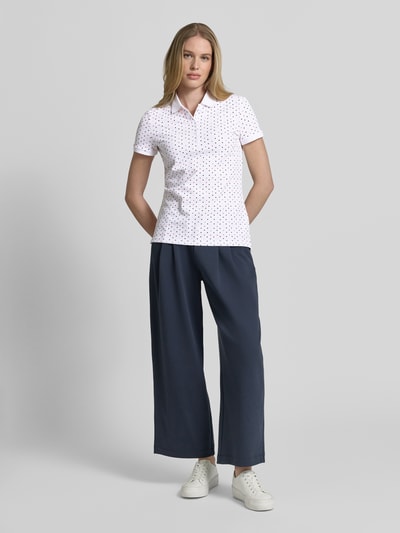 Modelka kobieca nosi Slim Fit Poloshirt mit Baumwoll-Anteil von Christian Berg Woman, wzór w ciemnoniebieskie kropki na białym tle, krój slim fit, bawełna z elastanem, kołnierzyk polo, krótkie rękawy, długość pleców w rozmiarze S: 61 cm, szerokie granatowe spodnie, białe sneakersy, pełny widok z przodu.