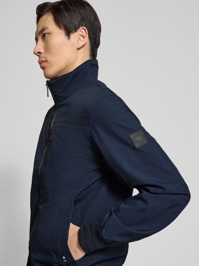 Männermodel trägt Comfort Fit Blouson mit geripptem Rumpfabschluss von Tom Tailor in Optik: Unifarben, Nahaufnahme Oberkörper. Passform: Comfort Fit, Kragenform: Stehkragen, Verschluss: Reißverschluss, Applikation: Logo-Patches, Details: Rückenpasse, Ärmelform: Langarm, Ärmelabschluss: Gerippt, Rumpfabschluss: Gerippt, Funktion: wasserabweisend, Taschen: Brustleistentasche, Napoleontasche, seitliche Eingrifftaschen, Materialverarbeitung: Stretch.