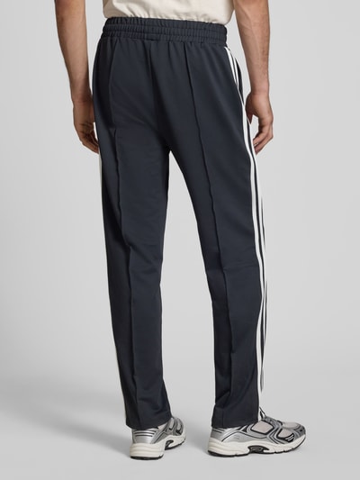 Męski model nosi spodnie dresowe Straight Fit Sweatpants mit Logo-Detail Modell 'Latucci' od Ellesse z elastycznej mieszanki materiałów, ozdobne szwy, elastyczny pas, czarne z białymi paskami, długość wewnętrzna nogawki w rozmiarze M: 79 cm, widok całej sylwetki od tyłu.