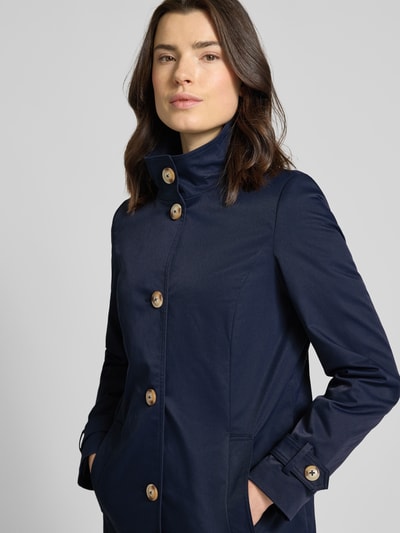 Frauenmodel trägt Trenchcoat mit Stehkragen und Ärmelriegeln von Christian Berg Woman Selection in Dunkelblau, Nahaufnahme Oberkörper. A-Linie, Knopfleiste, glatte Textur.
