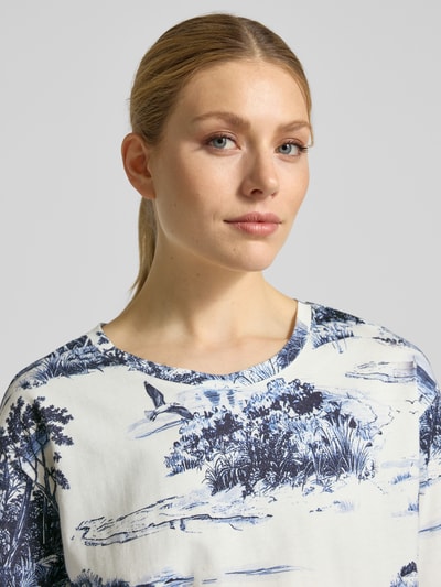 Frauenmodel trägt T-Shirt im Allover-Look Modell 'Hamptons Landschaft' von monari in Weiß mit blauem Landschaftsmuster, Nahaufnahme Oberkörper.