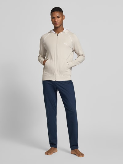 Männermodel trägt Regular Fit Sweatjacke aus Baumwoll-Mix von BOSS in Beige, Kapuze, Kontraststreifen, Kängurutasche, Ganzkörperansicht von vorne.