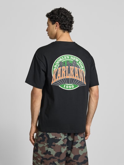 Männermodel trägt Oversized T-Shirt mit Logo-Stitching von KARL KANI aus reiner Baumwolle, Schwarz mit buntem Rückendruck, Ganzkörperansicht Rückansicht.